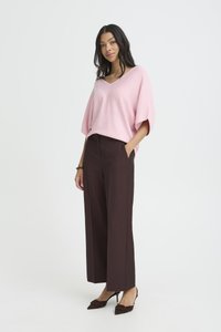 Frau steht in weitem, dunkelbraunem Hosenrock, lockerem rosa V-Ausschnitt-Oberteil und braunen spitzen Pumps vor einem schlichten weißen Hintergrund.