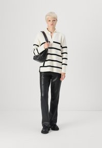 Sweter w kremowo-czarną paski z kołnierzykiem, zestawiony z czarnymi spodniami z ekologicznej skóry i czarną torebką na ramię. Buty są czarne.