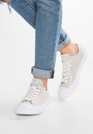 Lichtbeige lage sneakers met witte veters en zolen, gedragen met opgerolde blauwe jeans op gekruiste benen tegen een witte achtergrond.