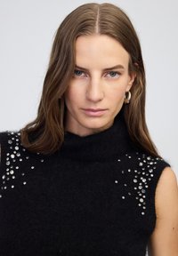 Czarny bezrękawnik z golfem, wykonany z miękkiego materiału, ozdobiony srebrnymi cyrkoniami ułożonymi w wzór na ramionach.