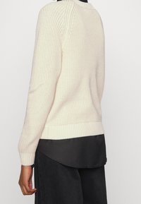 Kraftig stickad krämfärgad tröja med ribbade manschetter och nederkant, lagd över en svart topp. Har en klassisk rund halsringning och textil med struktur.