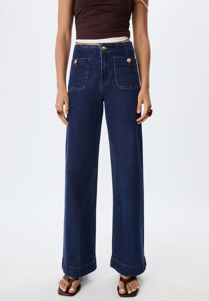 Femme portant un jean bleu foncé taille haute à jambes larges avec de grandes poches devant, un haut marron sans manches et des sandales marron.