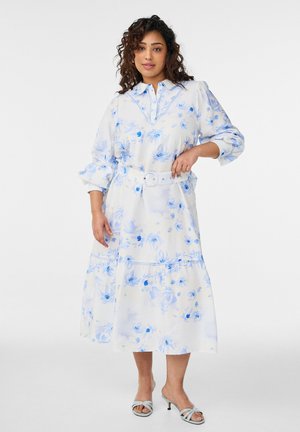 Žena oblečená v bielej midi šaty s modrými kvetinovými vzormi, opaskom v páse, dlhými rukávmi a svetlomodrými otvorenými sandálmi, stojaca na bielom pozadí.