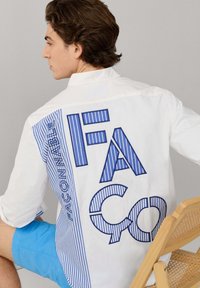 Jonge man zittend op een houten stoel, met een wit shirt waarop de blauwe gestreepte tekst "FACCNA" op de achterkant staat en felblauwe korte broek.