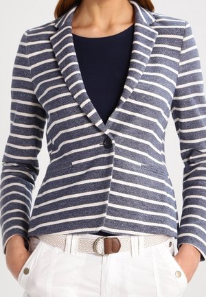 Kvinde iført en tætsiddende navy- og hvide vandrette stribet blazer over en navy skjorte, kombineret med hvide bukser og et beige bælte med en brun spænde.