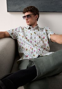 Chemise blanche à manches courtes avec un motif floral multicolore. Le modèle porte des lunettes de soleil, assis sur un canapé gris, avec un pantalon vert.