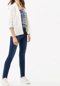 Cárdigan blanco de canalé sobre una blusa azul con estampado, combinado con jeans azul oscuro y zapatillas blancas. Un atuendo simple y casual.