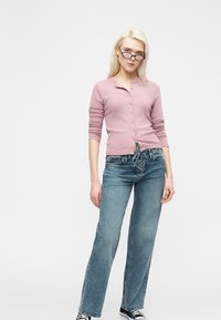 Jeune femme portant un haut rose à boutons, un jean bleu taille haute à lacets, des baskets noires et des lunettes, debout devant un fond clair uni.