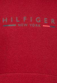 Red sweatshirt with a smooth texture, featuring a prominent printed "HILFIGER NEW YORK" label in gray and small colored accents below.