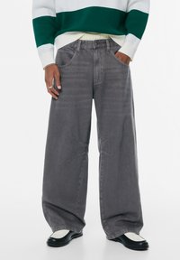Hellegraue Jeans mit weitem Bein und leicht abgenutzter Textur. Verfügt über Vordertaschen und einzigartige Kreuzstichdetails. Kombiniert mit schwarzen Schuhen.