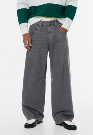 Breda grå denimbyxor med en lätt sliten textur. Har framfickor och unika korsstygnsdetaljer. Matchas med svarta skor.