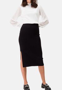 Femme portant un haut blanc à manches longues avec des manches transparentes rayées et une jupe noire midi avec une fente sur le côté, accompagnée de mocassins noirs.