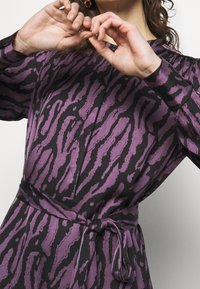 Lila och svart mönstrad klänning med långa ärmar, knyt i midjan och en mjuk tyg med en vågig, abstrakt tryck.