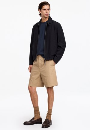 Jeune homme portant une veste noire zippée, une chemise bleu marine, un short beige jusqu'aux genoux, des chaussettes marron et des mocassins marron foncé, debout sur fond blanc.