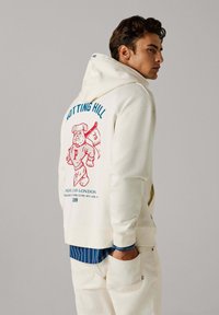 Off-white hoodie met een trekkoord, met een rode afbeelding van een beer en de tekst "NOTTING HILL". Gestyled over een blauw gestreept shirt.