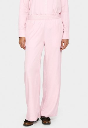 Persoon die losse lichtroze broek met elastische taille en bruine schoenen draagt, staand tegen een effen lichte achtergrond.
