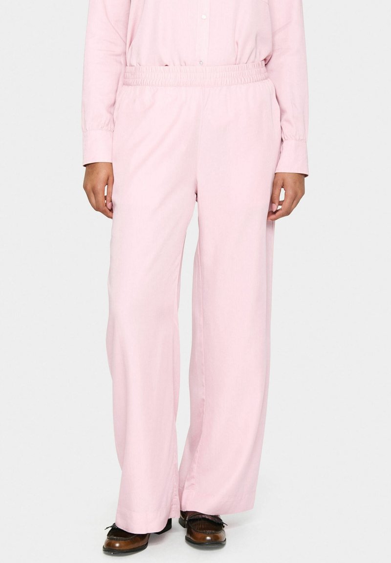 Personne portant un pantalon ample rose clair avec une taille élastique et des chaussures marron, debout devant un fond clair uni.