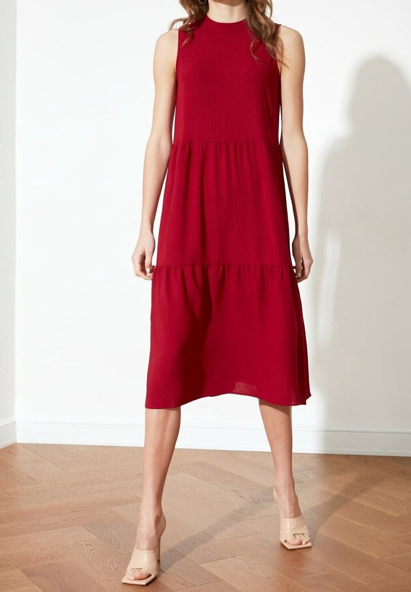 Femme portant une robe midi rouge sans manches et des talons beiges ouverts, debout sur un sol en bois contre un mur blanc.