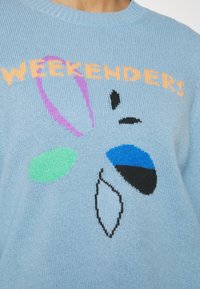 Pull en tricot bleu clair avec un design graphique coloré comportant le mot "WEEKENDERS" en orange, et des formes abstraites en violet, vert, noir et bleu.