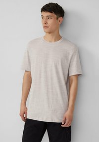 Jasny beżowy t-shirt z krótkim rękawem, okrągłym dekoltem, gładką teksturą i minimalnym poziomym wzorem. Noszony z czarnymi spodniami.