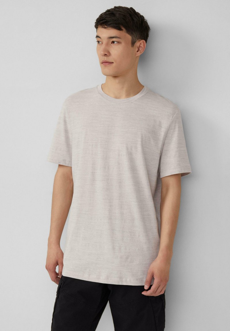 Jasny beżowy t-shirt z krótkim rękawem, okrągłym dekoltem, gładką teksturą i minimalnym poziomym wzorem. Noszony z czarnymi spodniami.