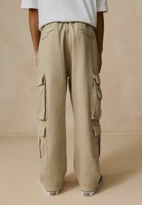 Pantalon cargo beige avec taille élastique, poches latérales et poches à rabat superposées. Le tissu est doux avec une coupe décontractée et ample, agrémentée de coutures.