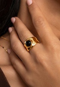 Bague en or avec un design lisse et ondulé, mettant en avant une pierre noire centrale. La bague a une finition polie et est portée sur une main avec des ongles bien entretenus.