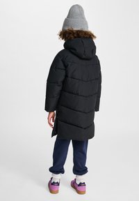 Sort pufferjakke med hætte, der har et chevron-quiltet design. Båret over marineblå bukser og lilla sko. Grå strikket hue tilføjer varme.