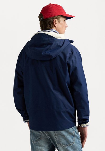 Marineblå vandafvisende jakke med hætte, med en afslappet pasform og hvidt indvendigt foer, parret med lettere slidte blå jeans og en rød kasket.