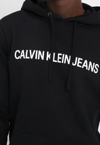 Sudadera negra con una capucha espaciosa, cordones y texto blanco en negrita "CALVIN KLEIN JEANS" en el pecho. Fabricada con tela suave.