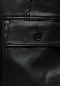 Μαύρο δερμάτινο ένδυμα με ραμμένη τσέπη με καπάκι, ασφαλισμένη με ένα μαύρο κουμπί, που δείχνει λεπτομερή υφή και ραφές.