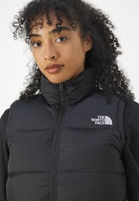 Μαύρο αδιάβροχο γιλέκο με ψηλό γιακά, μπροστινό φερμουάρ και λογότυπο εμφανές στην αριστερή πλευρά. Λείο και ραμμένο υφή με εφαρμοστή σχεδίαση.