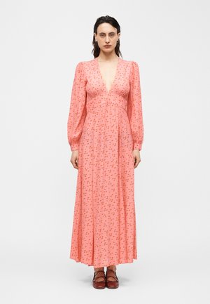 Kobieta w długiej różowej sukience z drobnym pomarańczowym kwiatowym wzorem, z głębokim dekoltem w serek, długimi rękawami i bordowymi sznurowanymi butami, stojąca na białym tle.