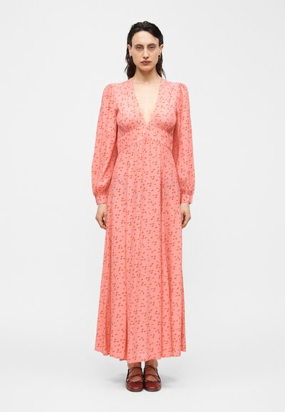 Mujer con un vestido largo rosa con pequeños estampados florales naranjas, escote en V profundo, mangas largas y zapatos burdeos con cordones, de pie contra un fondo blanco.