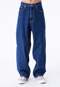Lockere blaue Jeans aus Denim mit hoher Taille, fünf Taschen und kontrastierenden Nähten. Kombiniert mit hellen Leinenschuhen.