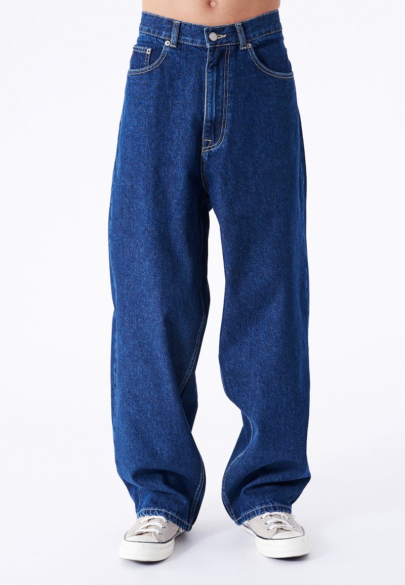 Lockere blaue Jeans aus Denim mit hoher Taille, fünf Taschen und kontrastierenden Nähten. Kombiniert mit hellen Leinenschuhen.