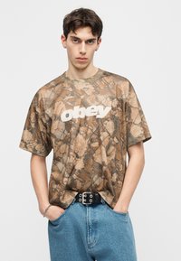 Νέος άνδρας που φοράει καφέ μπλουζάκι Obey με σχέδιο φύλλα, μπλε τζιν, μαύρη ζώνη, στέκεται με τα χέρια στις τσέπες μπροστά σε απλό φόντο.