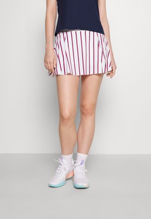 Personne portant une jupe plissée blanche avec des rayures verticales rouges, des chaussettes blanches, des baskets blanches avec semelles bleues et un haut noir, debout contre un mur blanc.