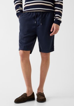 Homme portant un short bleu marine à cordon, une chemise rayée à manches longues et des mocassins en daim marron, debout les mains dans les poches.