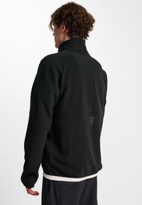 Svart fleecejacka med hög krage, långa ärmar och textilstruktur. Har en minimalistisk design med broderade detaljer på baksidan.
