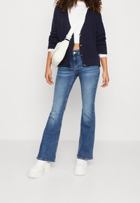 Cardigan blu navy a maglia con bottoni, canottiera bianca aderente, jeans blu svasati e stivali platform bianchi chunky, creando un look informale a strati.