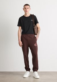 Melnas kokvilnas T-krekls, kas kombinēts ar tumši bordo sporta biksēm ar logo. Modelis valkā baltas kedas un brūnu pleca somu.