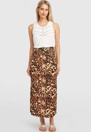 Femme portant un chemisier blanc sans manches et une longue jupe noire à motif feuilles tropicales beige, debout devant un fond clair uni.