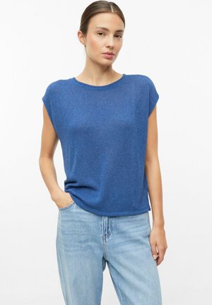Vrouw met bruin haar die een blauwe mouwloze gebreide top en lichtblauwe spijkerbroek draagt, staat met één hand in haar zak tegen een witte achtergrond.