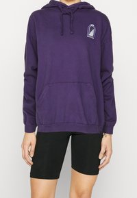 Lila hoodie i bomull, med en framlomme och en grafisk design på vänster bröstområde. Långa ärmar med ribbade muddar.