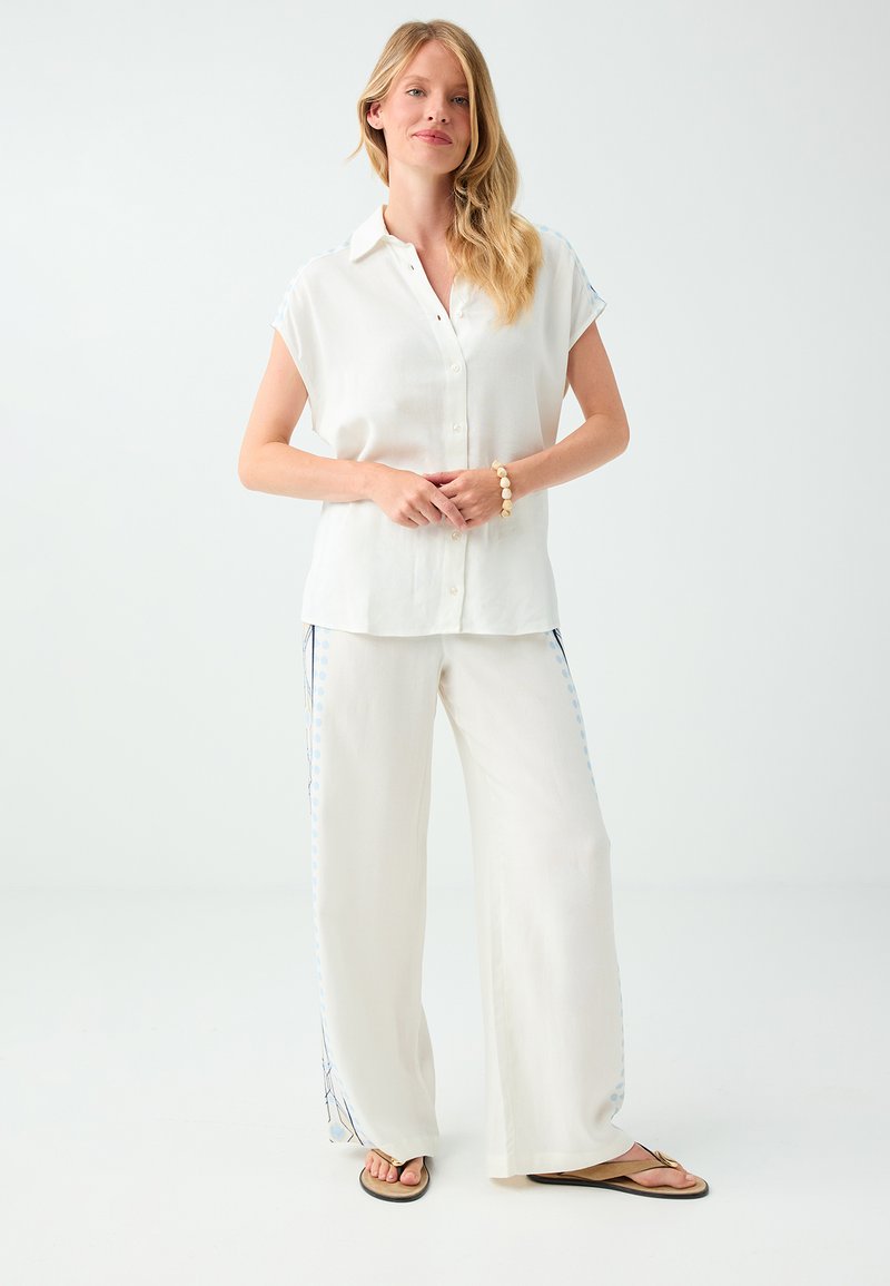 Femme portant une chemise blanche ample à manches courtes boutonnée, un pantalon blanc à jambes larges avec des accents bleus, des sandales beiges et un bracelet de perles.