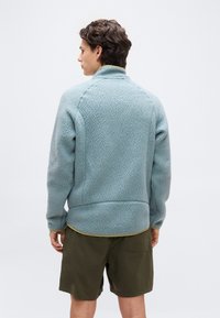 Άνδρας με πλάτη φοράει ανοιχτό μπλε πλαστική μπουφάν με υφή και χακί σορτς σε απλό λευκό φόντο.