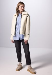 Giacca in finta pelliccia crema con rifiniture nere, indossata sopra una camicia a righe azzurre chiare, abbinata a jeans scuri e scarpe con zeppa beige.