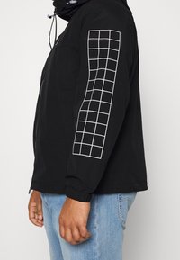 Svart jacka med dragkedja fram, elastiska ärmslut och ett rutmönster på den vänstra ärmen. Tillverkad av ett mjukt, lätt material.