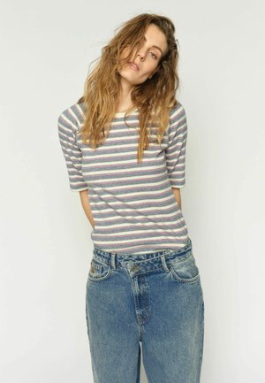 Noor naine, kellel on pikad lainelised juuksed, kannab lühikeste varrukatega triibulist särki ja lahtisi kõrge vöökohaga siniseid teksapükse tavalise tausta ees.